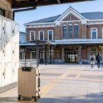福島県の地方駅をイメージした落ち着いたレンガ調の駅舎を背景に、旅行用キャリーケースと清潔なコインロッカーを自然に配置