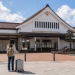 福島県の会津若松駅をイメージした伝統的な白い駅舎の正面、駅前広場に立つ旅行者と中型のキャリーケース、駅舎の屋根下に自然に配置されたコインロッカー